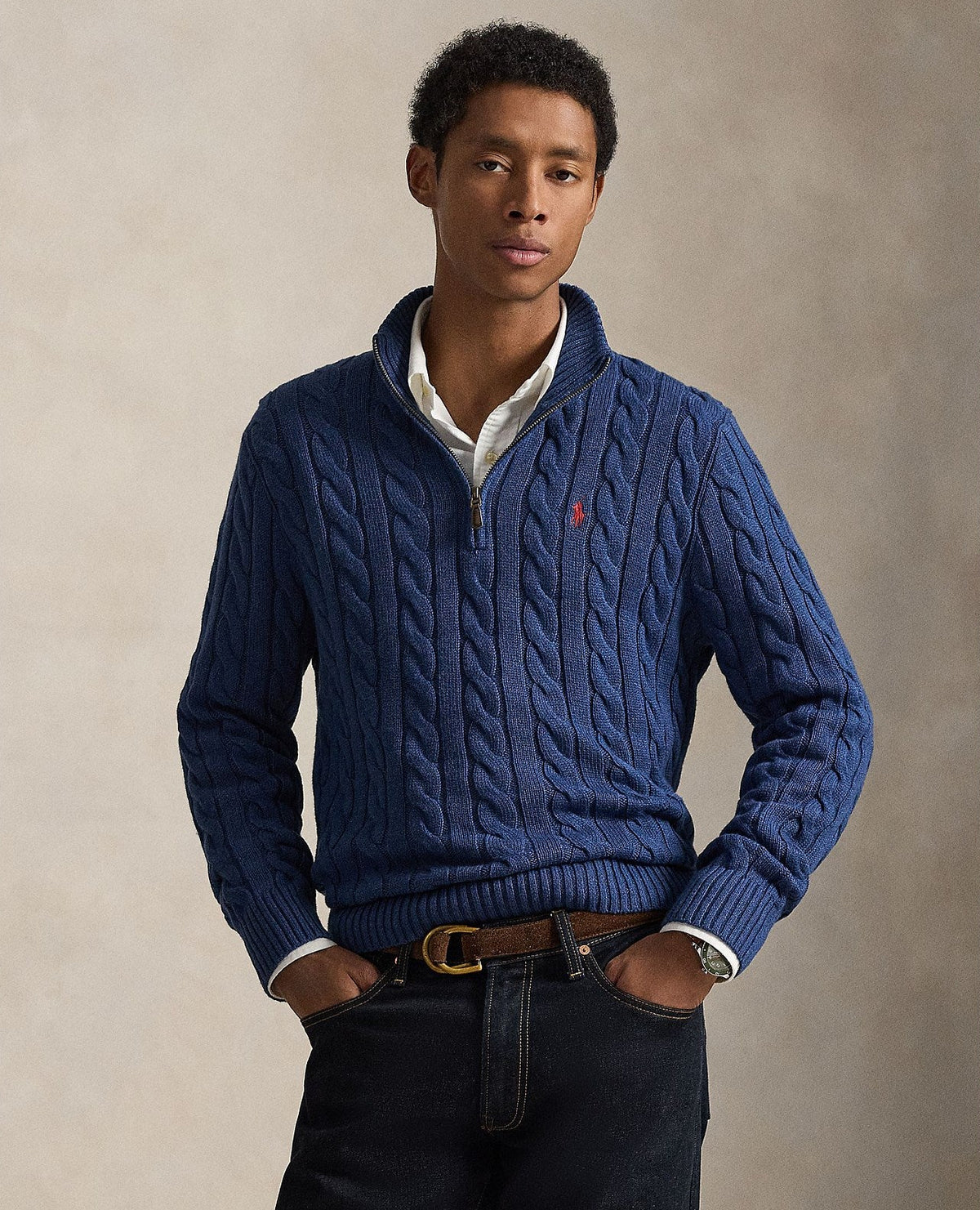 A man wearing a navy blue cable-knit quarter-zip sweater with a small embroidered polo player logo on the chest, styled with a white collared shirt, dark jeans, and a brown belt, set against a neutral beige studio background.