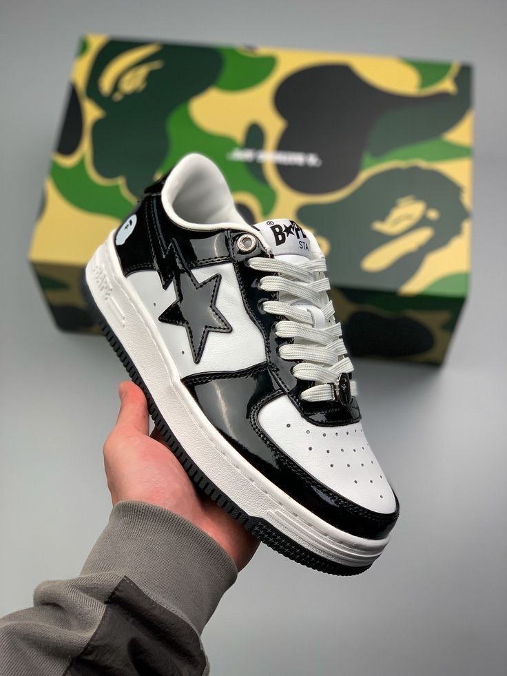 A hand holds a black and white BAPE sneaker in front of its green and black camouflage box.